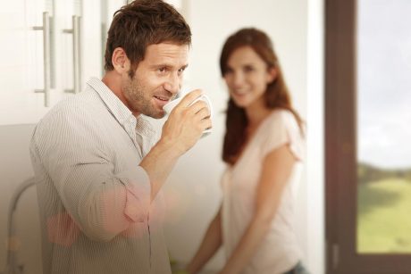 Mann mit Kaffeebecher lächelt zufrieden, im Hintergrund ein Holzfenster